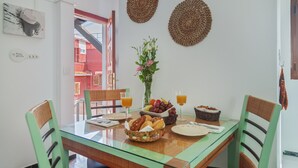 Un comedor con mesa de vidrio, sillas de mimbre, un florero con flores, fruta y vasos de jugo.