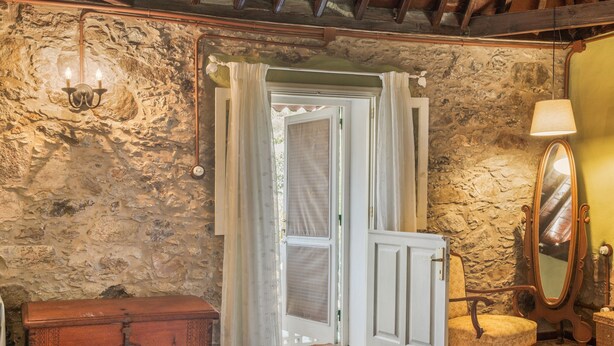 Una habitación rústica con pared de piedra, un cofre de madera, una silla, un espejo y una puerta con cortinas.