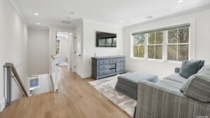 Amplia sala de estar con un sofá gris, suelo de madera, cómoda gris y un ventanal con vistas a árboles.