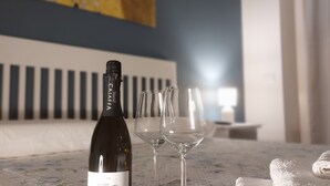 Une bouteille de vin et deux verres sur une table, dans une pièce aux murs bleus, avec un tableau accroché au-dessus du lit.