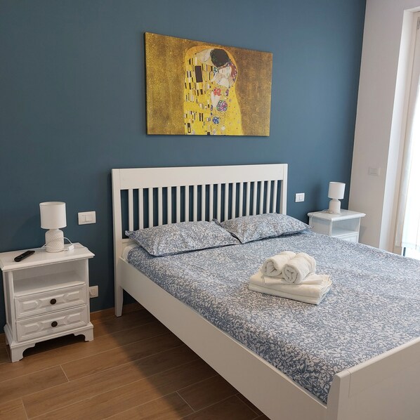 Une chambre avec un lit blanc, une table de chevet avec une lampe, un tableau accroché au mur et une serviette pliée posée sur le lit.