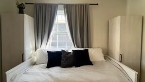 Un dormitorio con una cama grande, una ventana con cortinas, una mesita de noche y una planta pequeña.