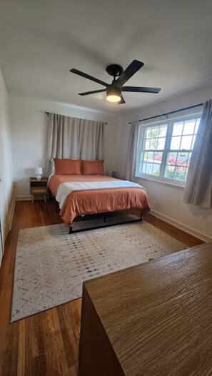 Um quarto com cama, ventilador de teto, mesa de cabeceira, janela com cortinas e piso de madeira.