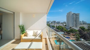 Un balcón con un sofá de mimbre y una mesita pequeña, con vistas a una piscina y al paisaje urbano.