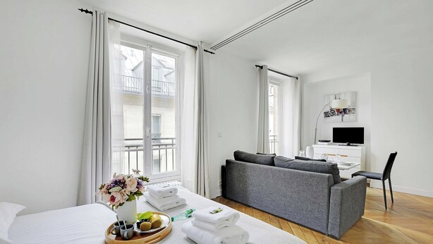 A hotel room with a bed, a grey sofa, a TV, and a dining area with a tray of fruits and towels. - Authentic Apartment - 1br/2p - Palais Garnier (Paris)