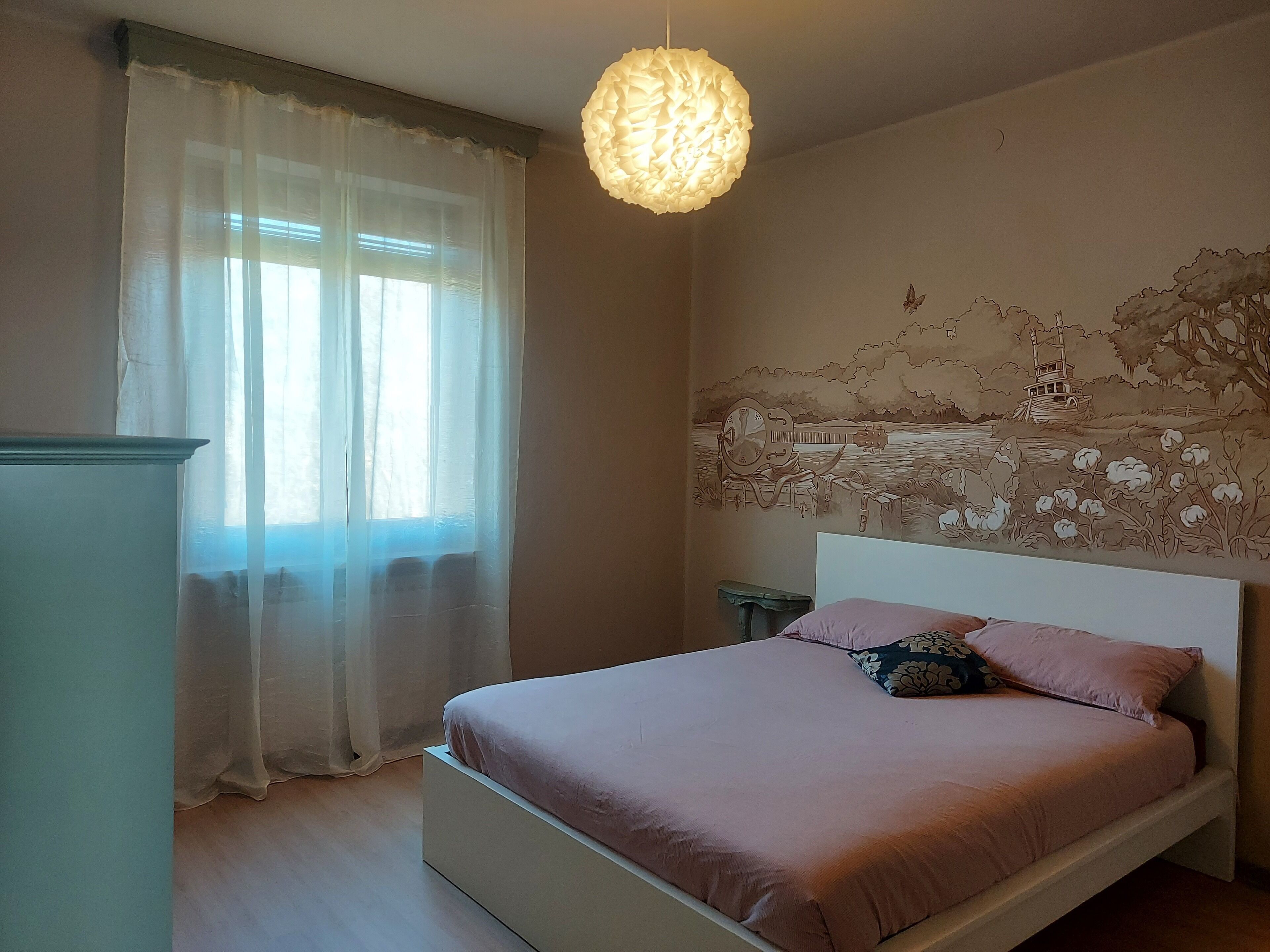 Ein Schlafzimmer mit einem Bett, einem Fenster mit transparenten Vorhängen, einer Wandmalerei und einer Pendelleuchte.