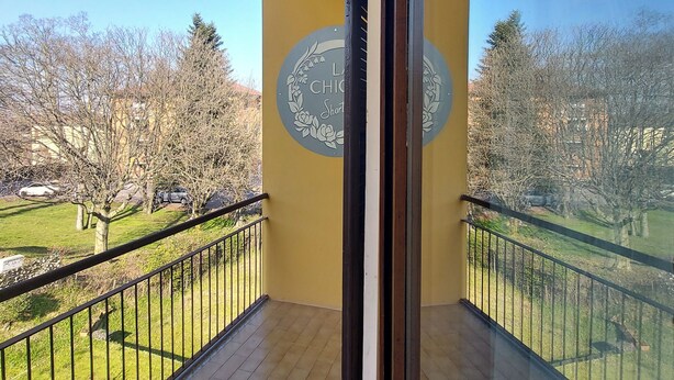 Un balcón con pared amarilla y puerta de cristal, con vistas a un parque con árboles y cielo despejado.