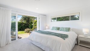 A bedroom with a large bed, white curtains, a window with a view of greenery, and a small bedside table with a lamp. - Whitewater - Stylish Beach House With Ocean Views (AVOCA BEACH)
