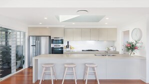 A modern kitchen with a central island, stainless steel appliances, and a large window. - Whitewater - Stylish Beach House With Ocean Views (AVOCA BEACH)