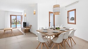 Un espace repas moderne avec une table en bois dressée pour six personnes, des chaises blanches et une grande fenêtre donnant sur une piscine et des montagnes.