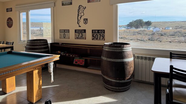 A pool table with a wooden frame and a green felt surface, two wooden barrels, a billiards cue, and a view of a rural landscape through large windows. - Posada Ruta Nacional 40  (Cabo Virgenes)