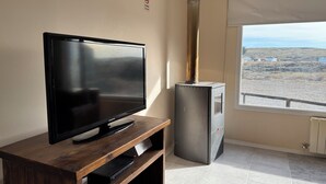 A room with a flat-screen TV on a wooden stand, a built-in fireplace, and a view of a rural landscape through a large window. - Posada Ruta Nacional 40  (Cabo Virgenes)