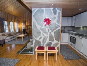 Un salon moderne avec un mur d’accent en pierre, une horloge rouge et un coin cuisine doté de meubles blancs.