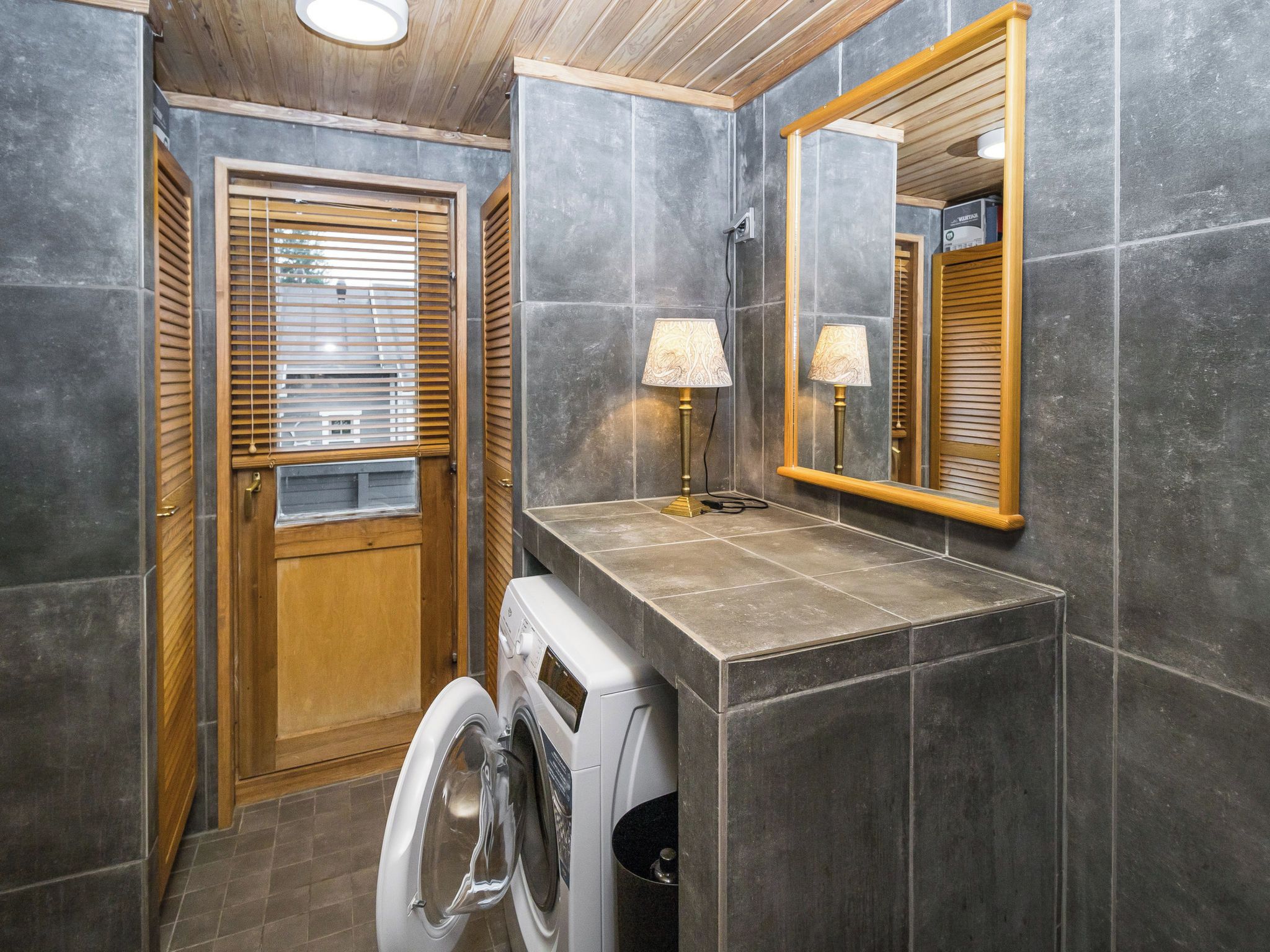 Een kleine badkamer met een wasmachine, houten jaloezieën en een spiegel.