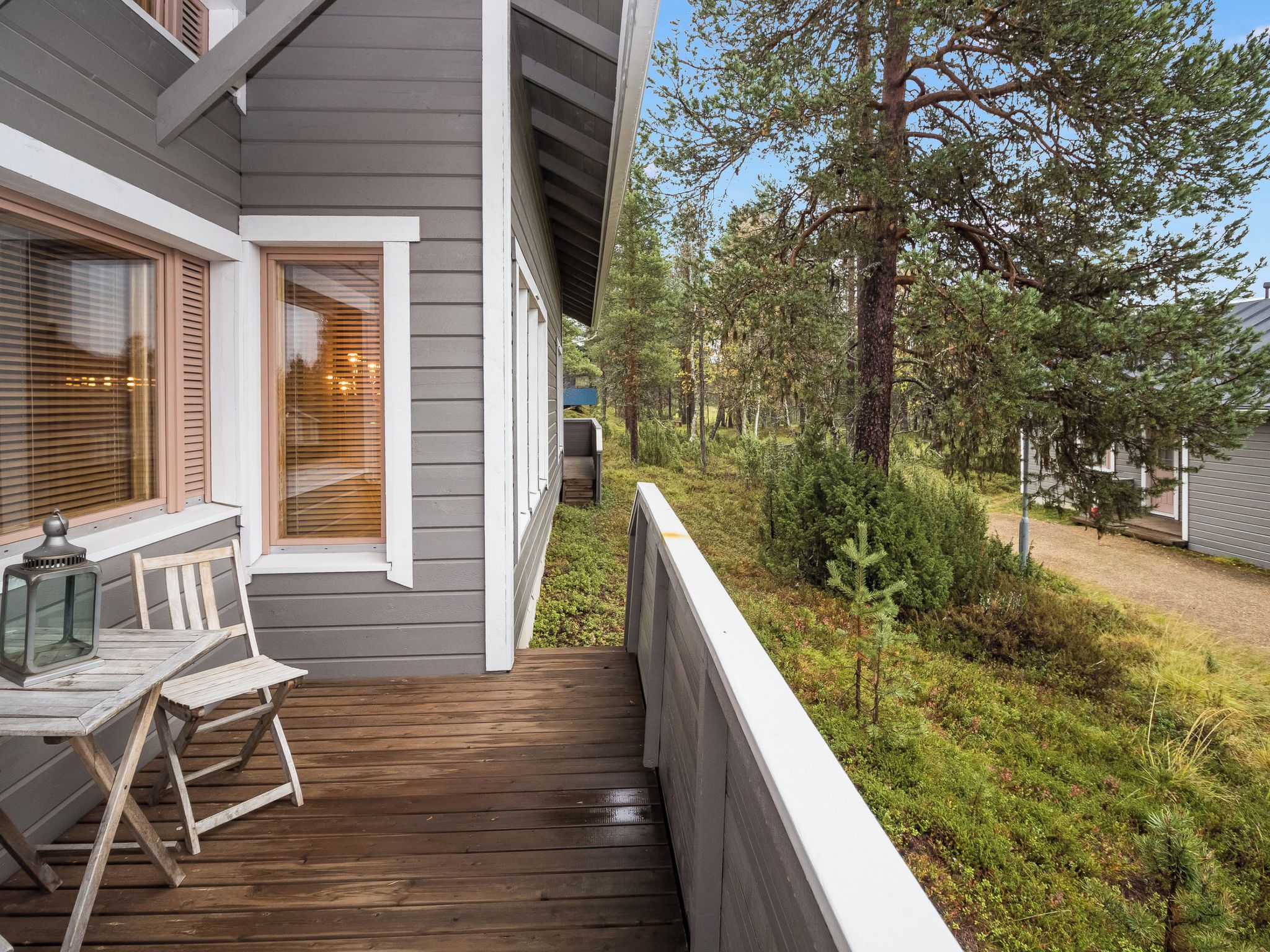 A wooden deck with a table and chairs, a window with blinds, and a view of a forested area.