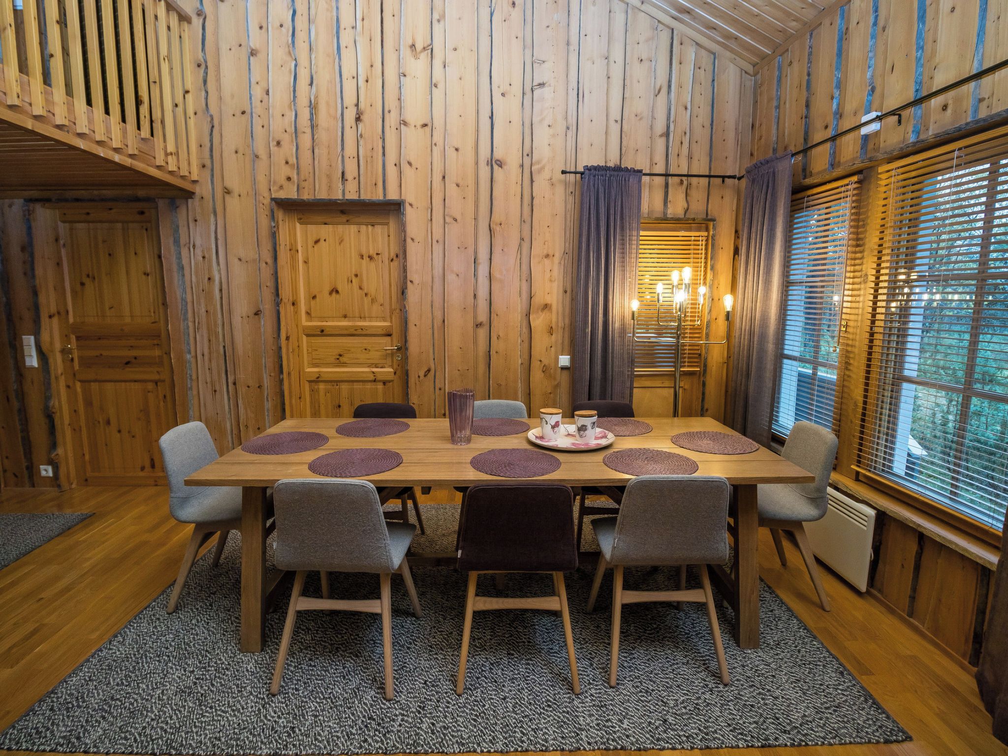 Een kamer met houten lambrisering, een grote tafel gedekt voor zes personen, omringd door stoelen, en een raam met jaloezieën.