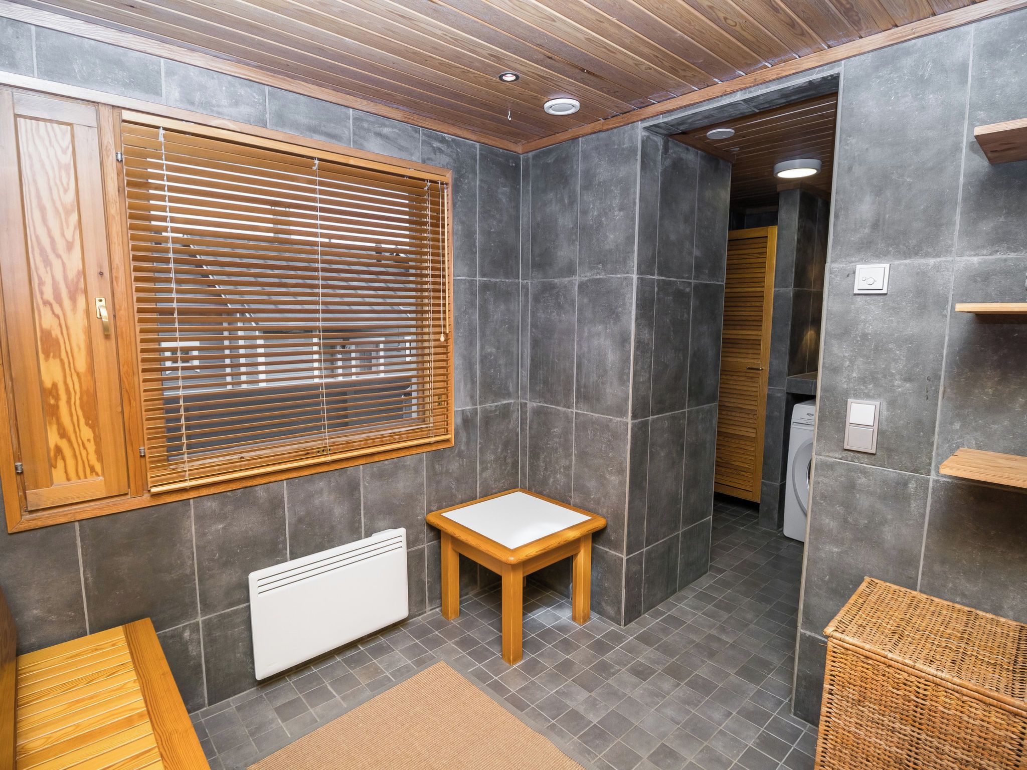 Un baño con piso de madera, un radiador, una mesita y un ventanal con persianas.