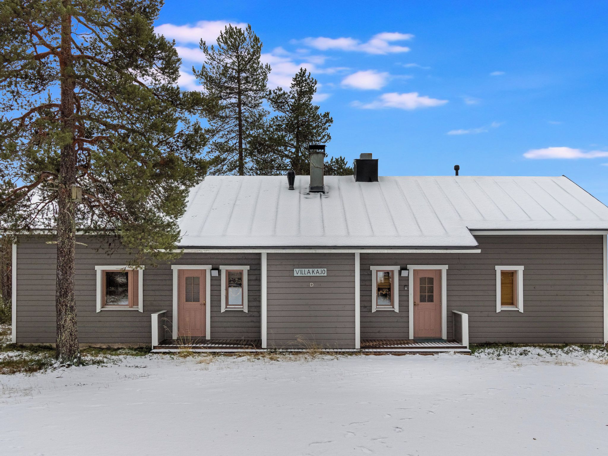 En tvåvåningsbyggnad med ett tak av metall, en skylt med texten ”VILLA KAJO” och en snötäckt mark.