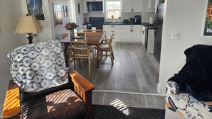 Een moderne keuken met witte kastjes, een eethoek met een houten tafel en stoelen, en een zithoek met een bank en een decoratief plaid.