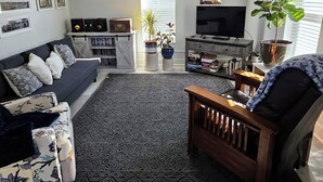 Een moderne woonkamer met een donker houten vloer, een zwarte bank met opvallende kussens, een houten salontafel, een televisie en een plafondventilator.