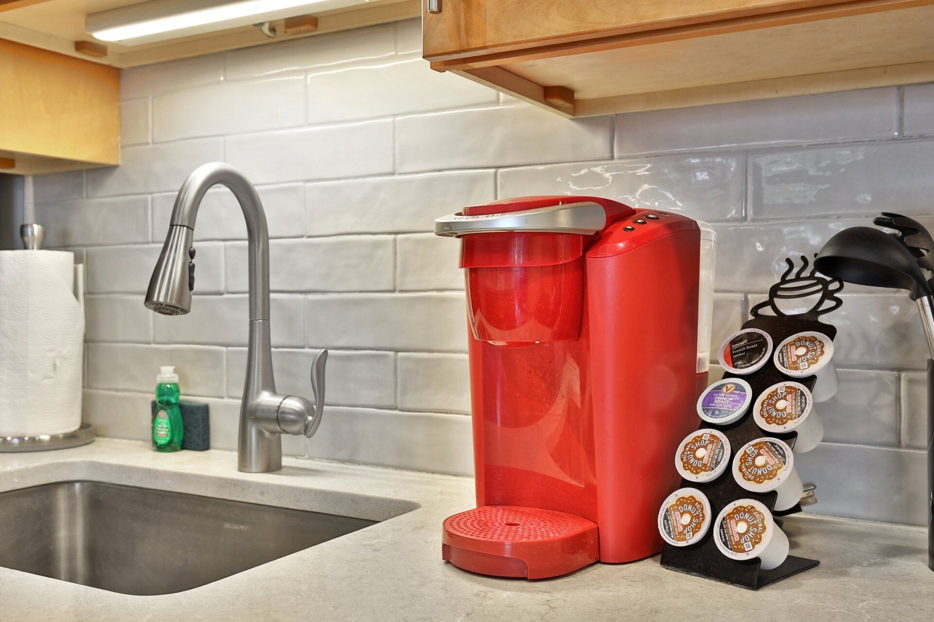 A kitchen with a stainless steel sink, a red coffee maker, and a coffee pod holder with multiple pods.