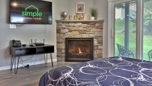A modern bedroom with a stone fireplace, a bed with a patterned duvet, a mounted TV, and a desk with a phone and router.