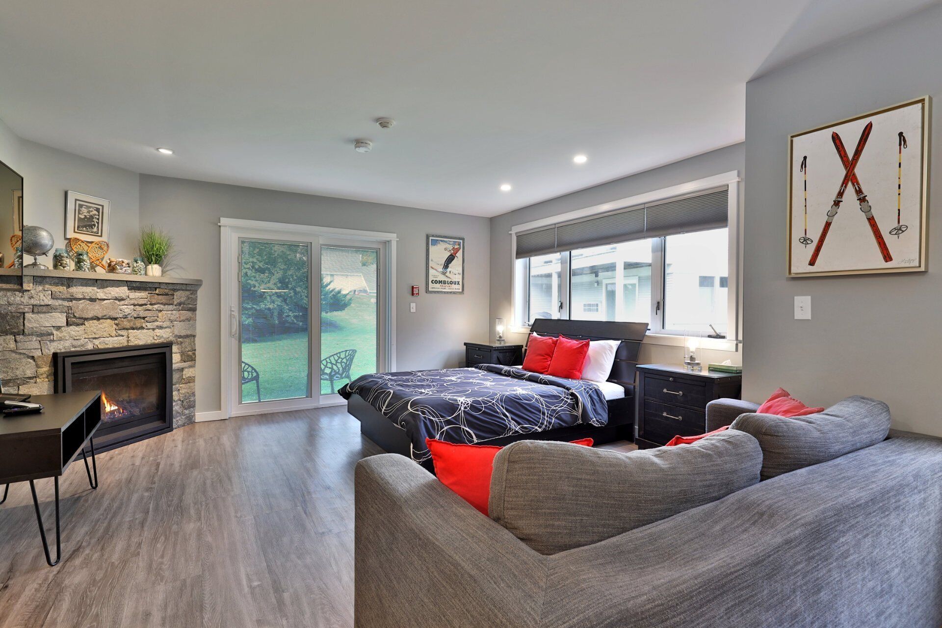 Un dormitorio moderno con chimenea, una cama grande con almohadas rojas y blancas, un sofá gris y un cuadro de esquíes en la pared.
