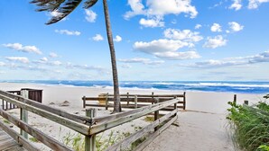 Ein hölzerner Steg führt zu einem Strand mit einer Palme und blauem, wolkenlosem Himmel.
