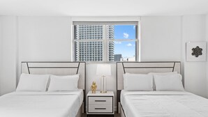 Ein Hotelzimmer mit zwei Betten, einem Nachttisch mit Lampe und Blick auf hohe Gebäude durch ein großes Fenster.