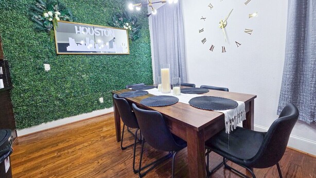Un coin repas avec une table en bois, des chaises noires, un mur vert orné d’une inscription « Houston » et une horloge murale.