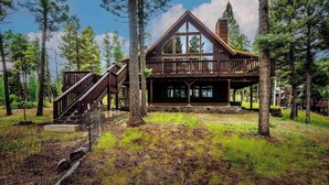 A cabin with a wooden deck, large windows, and a staircase leading to an upper level. - Spacious Mountain Cabin with Views, Deck, Second Living Area, New Hot Tub (Angel Fire)