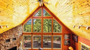 A cozy cabin interior with a stone fireplace, large windows offering a view of the deck, and wooden beams on the ceiling. - Spacious Mountain Cabin with Views, Deck, Second Living Area, New Hot Tub (Angel Fire)