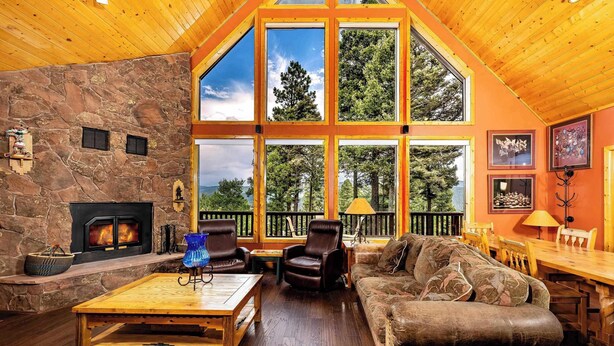 A spacious living room with a stone fireplace, leather furniture, large windows, and a wooden ceiling. - Spacious Mountain Cabin with Views, Deck, Second Living Area, New Hot Tub (Angel Fire)