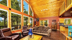 A spacious living room with leather sofas, a wooden coffee table, and large windows offering a view of trees and mountains. - Spacious Mountain Cabin with Views, Deck, Second Living Area, New Hot Tub (Angel Fire)