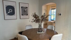 Un coin repas avec une table ronde en bois, quatre chaises grises à revêtement rembourré, un vase contenant des fleurs séchées et deux œuvres d’art abstraites encadrées accrochées au mur.