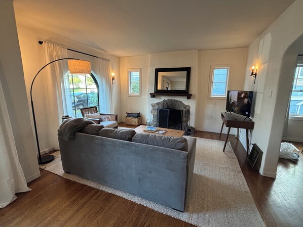 A living room with a grey sofa, a fireplace, a television, and a large window with curtains.