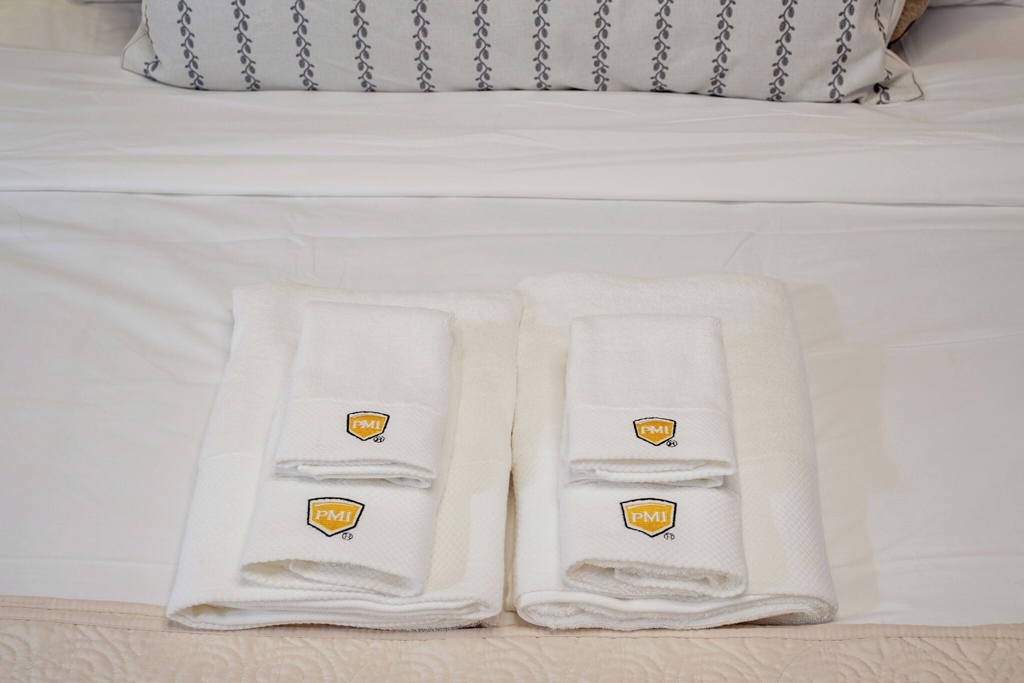 Un lit bien fait, avec du linge de lit blanc et deux serviettes pliées, chacune ornée d’un logo.
