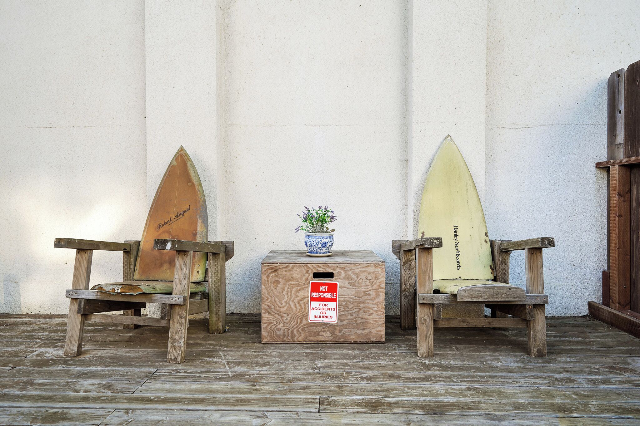 Deux chaises en bois ornées de planches de surf, une plante en pot et une boîte en bois sur laquelle est apposé un panneau d’avertissement.