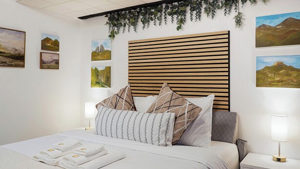 A neatly made bed with white linens and towels, a wooden slatted headboard, and wall-mounted artwork depicting landscapes. - Coast PalmTree Yard 2BR Block from Beach (Pacifica)