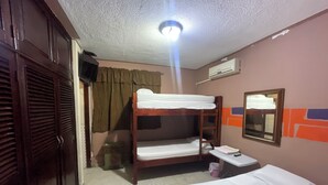 Habitación de hotel con dos literas, una cama individual, una mesita con un libro y un teléfono, un espejo y un equipo de aire acondicionado.