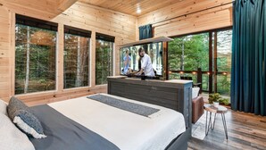 Un dormitorio moderno con una cama grande, televisión y vistas al bosque a través de amplios ventanales.