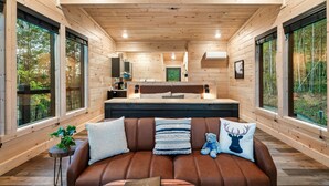 Interior de una cabaña de madera con una cama grande, un sofá de cuero marrón y vista al bosque a través de amplias ventanas.