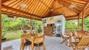 Un comedor al aire libre con mesas y sillas de madera, techo de paja y vistas a vegetación exuberante.