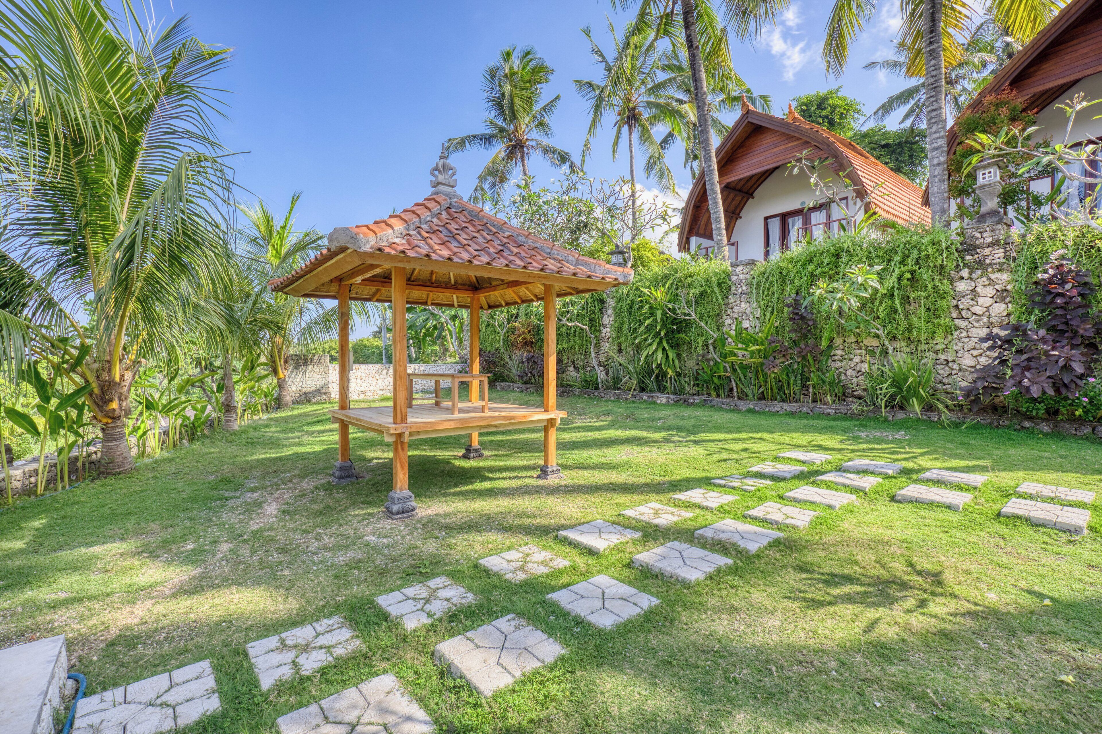 A gazebo in a well-maintained garden with stone pathways, surrounded by lush greenery and palm trees.