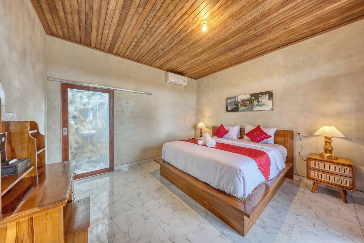 A bedroom with a wooden bed, a desk, a chair, a nightstand, and a wall-mounted air conditioner.