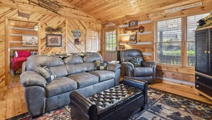 A cozy cabin interior with a leather sofa, a tufted ottoman, and a leather armchair. There are wooden walls, a fireplace, and a window with a view of greenery. - Lovey Bear - A Cozy Dog-Friendly Cabin with Hot Tub, Fireplace – Across from Soaky Mountain (Sevierville)