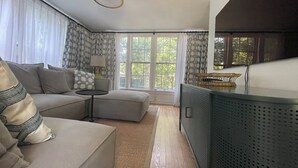 Una sala de estar moderna con sofá, reposapiés y una mesita. Hay ventanas grandes con cortinas, un radiador y una lámpara en el techo.