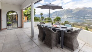 Un patio techado con mesa y sillas, vista a montañas y un lago, y una entrada con portón.