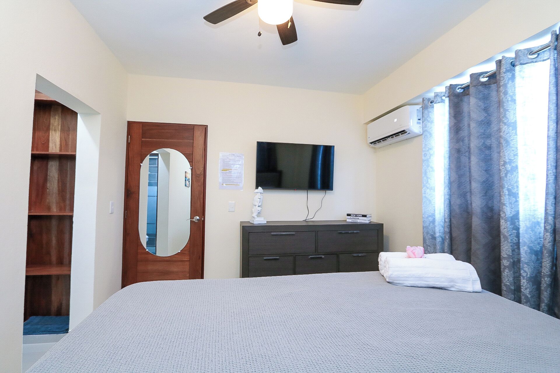Un dormitorio con cama, cómoda, espejo, televisor de pantalla plana y ventana con cortinas.
