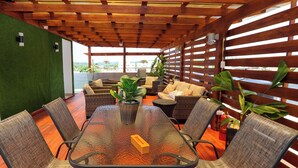 Un patio al aire libre con una mesa de vidrio rodeada de sillas de mimbre, plantas en macetas y una estructura de pérgola de madera.